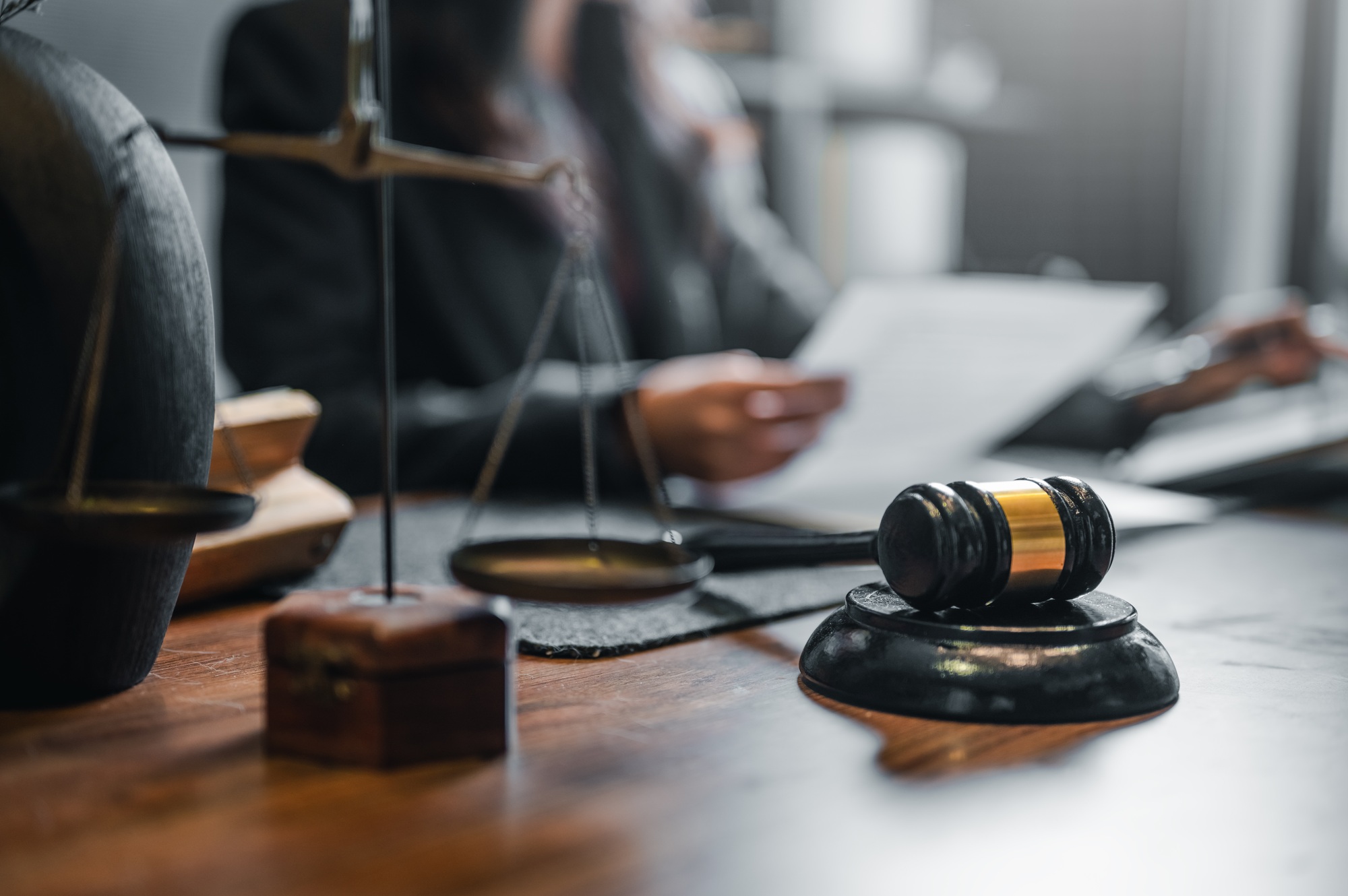 A judge's gavel sits on a wooden table next to a scale and a book. The scene suggests a courtroom.